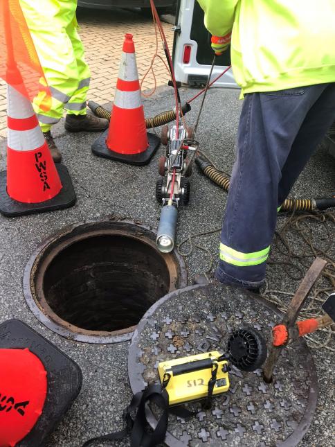 A small camera prepares to be lowered into a manhole to televise the condition of a sewer pipe.