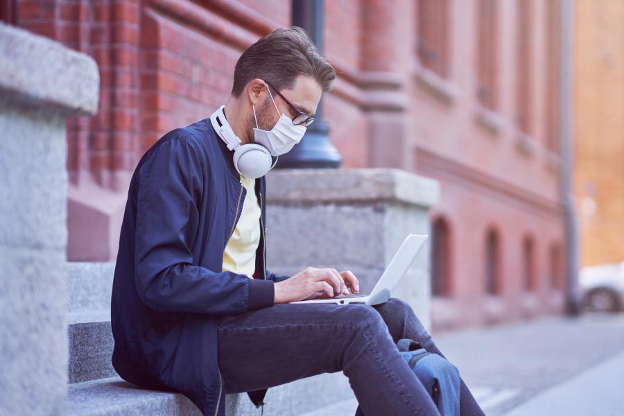 A college student wearing a mask sits on a building's steps typing on a laptop